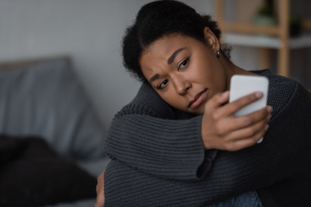 heartbroken multiracial woman in sweater using blurred smartphone in blurred bedroomの写真素材