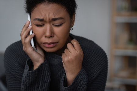 Crying multiracial woman talking on smartphone with helpline at homeの写真素材