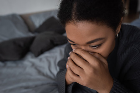 multiracial woman with mental problem covering face while sitting on bed at homeの写真素材