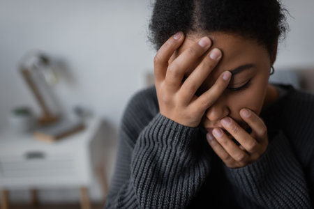 Young multiracial woman with depression touching face at blurred home