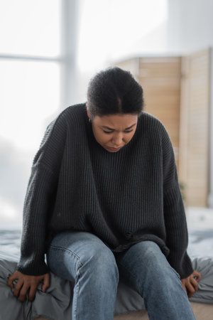 Young multiracial woman with mental problem sitting on bed at homeの写真素材