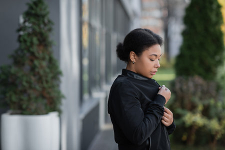 Upset multiracial woman with psychological problem wearing jacket on blurred urban street
