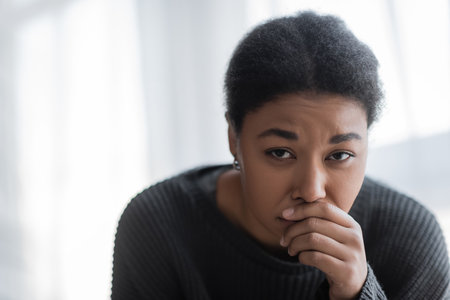 Displeased multiracial woman in knitted sweater looking at camera at home