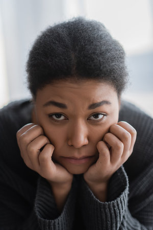 Depressed multiracial woman in sweater looking at camera at homeの写真素材