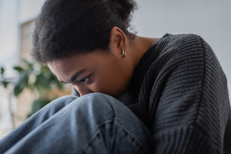 Portrait of frustrated multiracial woman in sweater and jeans sitting at homeの写真素材