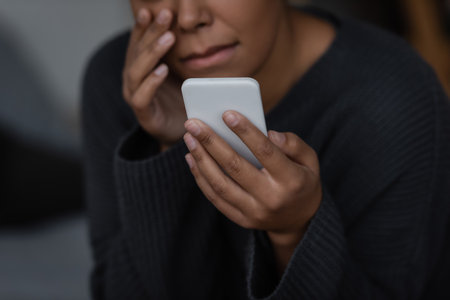 Cropped view of blurred multiracial woman with depression holding smartphone at homeの写真素材