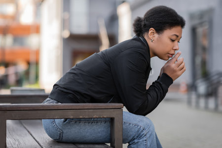 Disappointed multiracial woman in jacket sitting on bench on blurred urban streetの写真素材