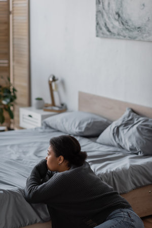 Young multiracial woman with psychological problem looking away near bed at homeの写真素材