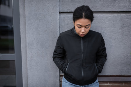 multiracial woman with mental problem looking down while standing near building on urban streetの写真素材