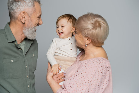 grandmother holding happy granddaughter near smiling bearded husband isolated on greyの写真素材