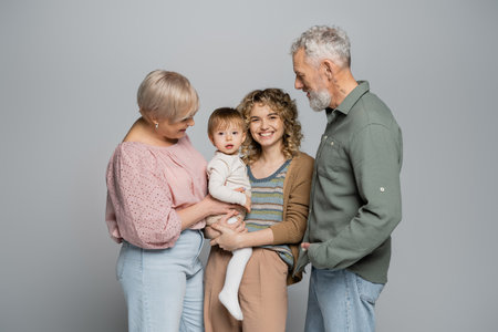senior couple looking at cheerful adult daughter with toddler child isolated on greyの写真素材