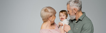 mature woman with bearded husband looking at cheerful toddler granddaughter isolated on grey, banner