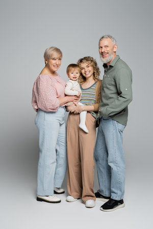 full length of happy middle aged grandparents and adult daughter with baby girl smiling at camera on grey backgroundの写真素材