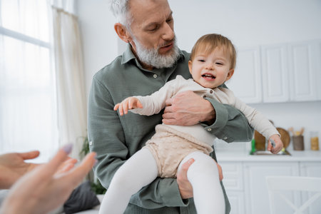 mature bearded man holding cheerful toddler granddaughter in kitchenの写真素材
