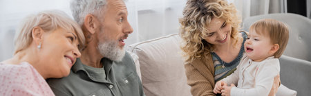 amazed bearded man looking at toddler granddaughter near family on couch in living room, bannerの写真素材