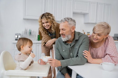 smiling bearded man holding bowl near granddaughter sitting on baby chair during breakfast in kitchenの写真素材