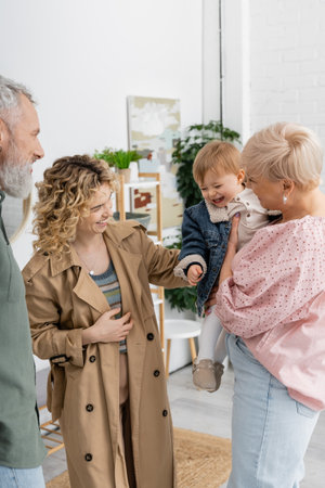 pleased woman in trench coat smiling near happy parents and little daughter at home