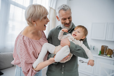cheerful middle aged couple having fun with smiling granddaughter holding spoon in kitchenの写真素材