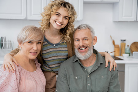 pleased middle aged couple smiling at camera near adult daughter in kitchen at homeの写真素材
