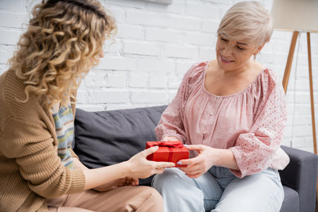 curly woman presenting gift to pleased mother while sitting on couch at homeの写真素材