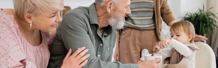 pleased grandparents looking at toddler child with spoon having breakfast in kitchen, bannerの写真素材