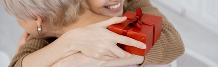 partial view of smiling woman with red gift box embracing middle aged mom at home, bannerの写真素材