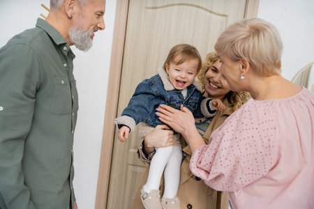 overjoyed child in denim jacket smiling near mother and grandparents near entrance door at home