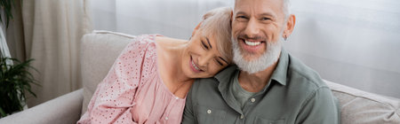 carefree middle aged man smiling at camera near happy wife on sofa in living room, banner