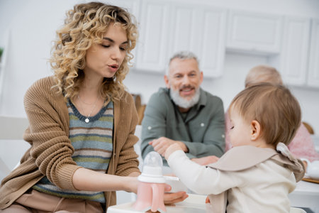 woman feeding little daughter with breakfast near mature parents smiling on blurred background in kitchenの写真素材