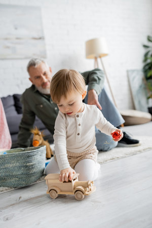 little girl playing with toy car on floor in living room near grandpa on blurred backgroundの写真素材