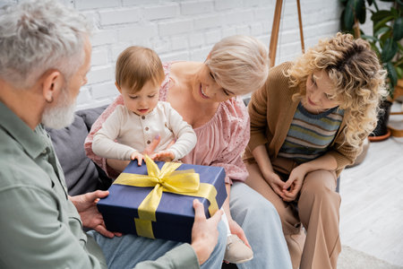 toddler child opening gift box near smiling mother and grandparents on couch in living roomの写真素材