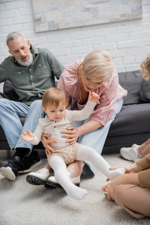 happy mature woman having fun with toddler granddaughter near family in living roomの写真素材