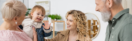 carefree girl waving hand near happy grandparents and smiling mother in living room, banner