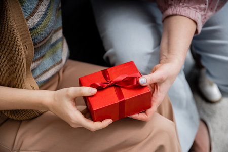partial view of woman holding red gift box near mature mother while sitting at homeの写真素材