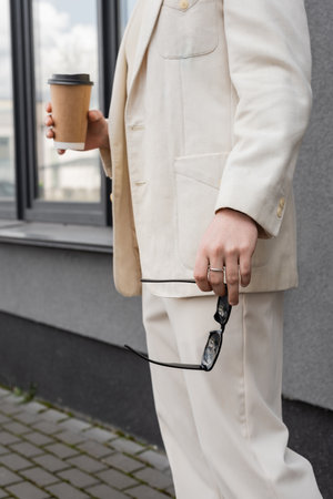 Cropped view of gay man in suit holding sunglasses and coffee to go outdoorsの写真素材