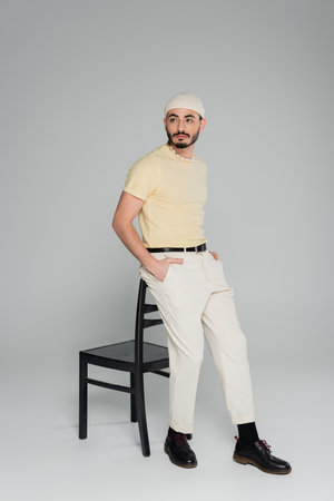 Young gay man in beige hat posing near chair on grey background