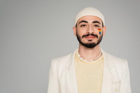 Portrait of stylish gay man with lgbt flag on cheek looking at camera isolated on greyの写真素材