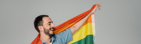 Overjoyed gay man holding lgbt flag and looking away isolated on grey, banner