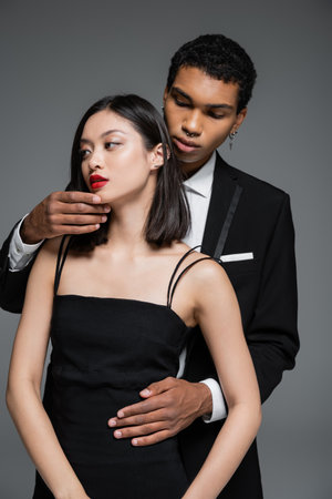 young african american man touching chin of elegant asian woman with red lips isolated on greyの写真素材