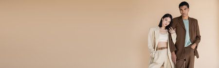 young interracial couple in trendy suits standing with hands in pockets isolated on beige, bannerの写真素材