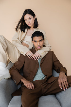 sensual asian woman embracing african american man in trendy suit sitting in armchair isolated on beigeの写真素材