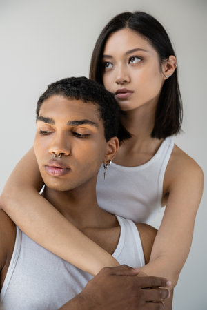 charming asian woman hugging young african american man with silver piercing isolated on greyの写真素材