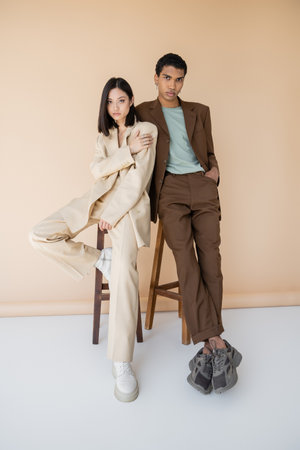 full length of young multiethnic couple in stylish suits posing near stools on beige backgroundの写真素材