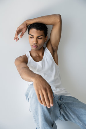 young african american guy in white tank top and jeans sitting on grey background