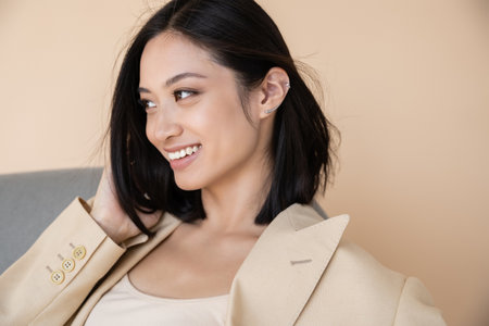 cheerful and stylish asian woman in blazer looking away on beige backgroundの写真素材