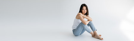 full length of slender asian model in blue jeans and white tank top sitting on grey background, bannerの写真素材
