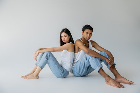 full length of barefoot and stylish interracial couple in jeans and tank tops sitting back to back on grey backgroundの写真素材