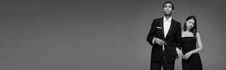 young interracial models in black elegant clothes looking at camera isolated on grey, banner