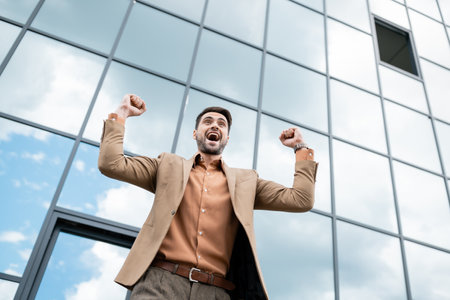 low angle view of joyful businessman screaming and showing success gesture near urban building with glass facadeの写真素材