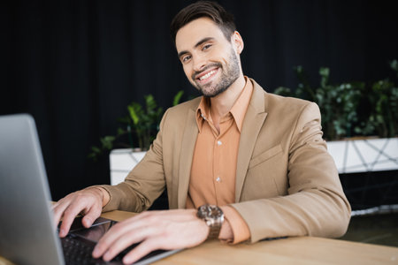 cheerful bearded businessman typing on laptop and looking at camera at workplace in officeの写真素材